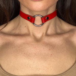 May include: A red leather choker necklace featuring a large silver ring. The choker fastens with a snap. This striking necklace is a bold accessory, designed to be worn around the neck.