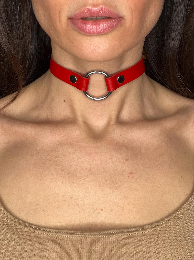 May include: A red leather choker necklace featuring a large silver ring. The choker fastens with a snap. This striking necklace is a bold accessory, designed to be worn around the neck.