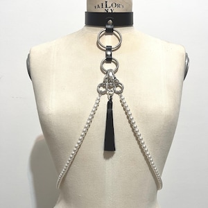May include: Black leather choker with silver-tone rings and a pearl strand harness. A black tassel hangs from the center. The choker has the text "TAILOR'S NY" printed on it.