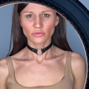 May include: A black leather choker necklace with a silver ring detail. The choker is worn around the neck. The model is wearing a beige vest top.
