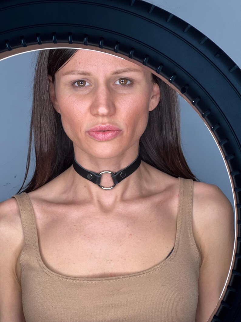May include: A black leather choker necklace with a silver ring detail. The choker is worn around the neck. The model is wearing a beige vest top.