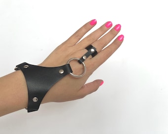 May include: Black leather wrist cuff with a ring attachment. The cuff is connected to a ring worn on the middle finger by a silver metal ring. The cuff is secured with silver rivets. The person's nails are painted a bright pink.