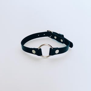May include: Black leather choker necklace featuring a large silver-coloured metal ring. The choker has a buckle fastening and silver-coloured rivets. The background is plain white.