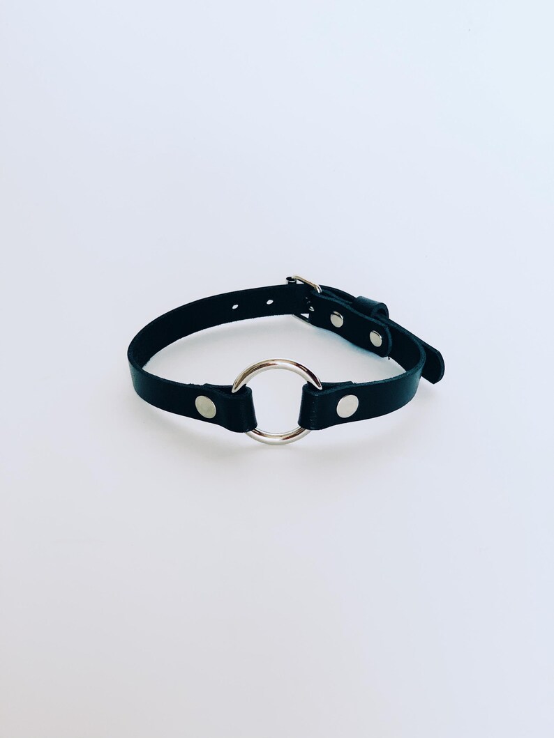 May include: Black leather choker necklace featuring a large silver-coloured metal ring. The choker has a buckle fastening and silver-coloured rivets. The background is plain white.