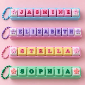 May include: Four rectangular keycap-style name keychains with ball chains. The top one is blue with pink letters spelling JASMINE. The second is pink with purple letters spelling ELIZABETH. The third is pink with yellow letters spelling STELLA. The bottom is green with green letters spelling SOPHIA. Each has a flower icon.
