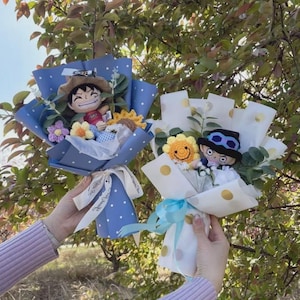 May include: Two bouquets featuring crocheted anime-style character dolls. One bouquet is wrapped in blue paper with white polka dots, the other in white paper with gold polka dots. Both include crocheted flowers, green foliage, and ribbons.