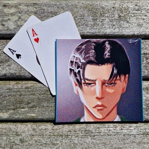 May include: Square art print with an anime-style portrait of a person with dark hair and a serious expression. Two playing cards, an Ace of Hearts and an Ace of Spades, are placed near the print.