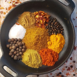 May include: A black cast iron pan filled with various spices, including salt, black peppercorns, allspice berries, turmeric, paprika, chili flakes, cumin, and coriander.