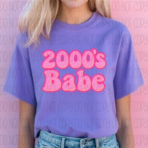 May include: A lilac t-shirt featuring the text "2000's Babe" in a pink, glittery font. The text is in a retro, bubble letter style. The shirt has short sleeves and a crew neck.