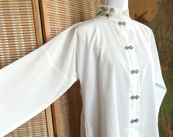 May include: A white blouse with a mandarin collar and decorative silver clasps. The collar is decorated with floral embroidery. The blouse has long sleeves and is paired with a beige skirt with a dragon design.