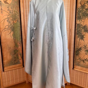 May include: A light blue, long-sleeved dress with a mandarin collar and a decorative clasp. The dress features a subtle floral pattern and side slits. The sleeves have tie closures. The dress is displayed on a white dress form.