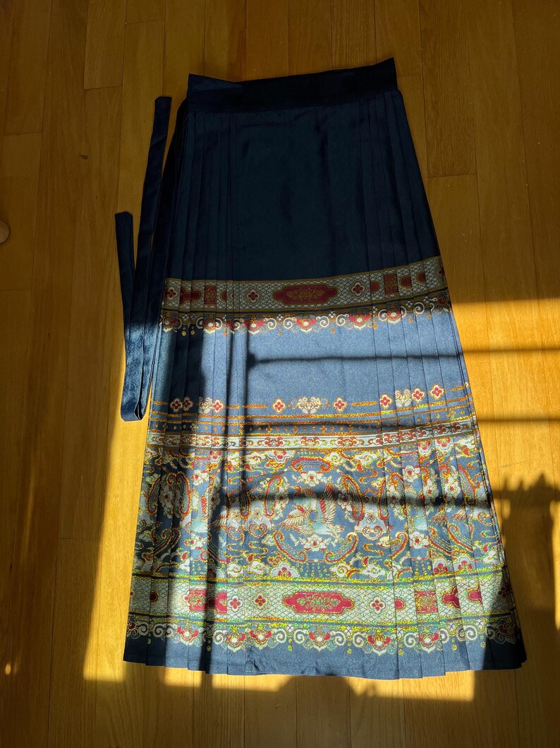 May include: A navy blue pleated skirt with a decorative patterned border. The border features red, yellow, and blue floral and paisley designs. The skirt has a matching navy blue tie.