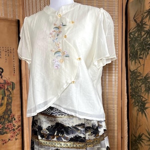 May include: Cream-coloured blouse with short sleeves and a mandarin collar. The blouse features an embroidered floral design and gold-coloured button closures. The blouse is paired with a patterned skirt with a landscape design.