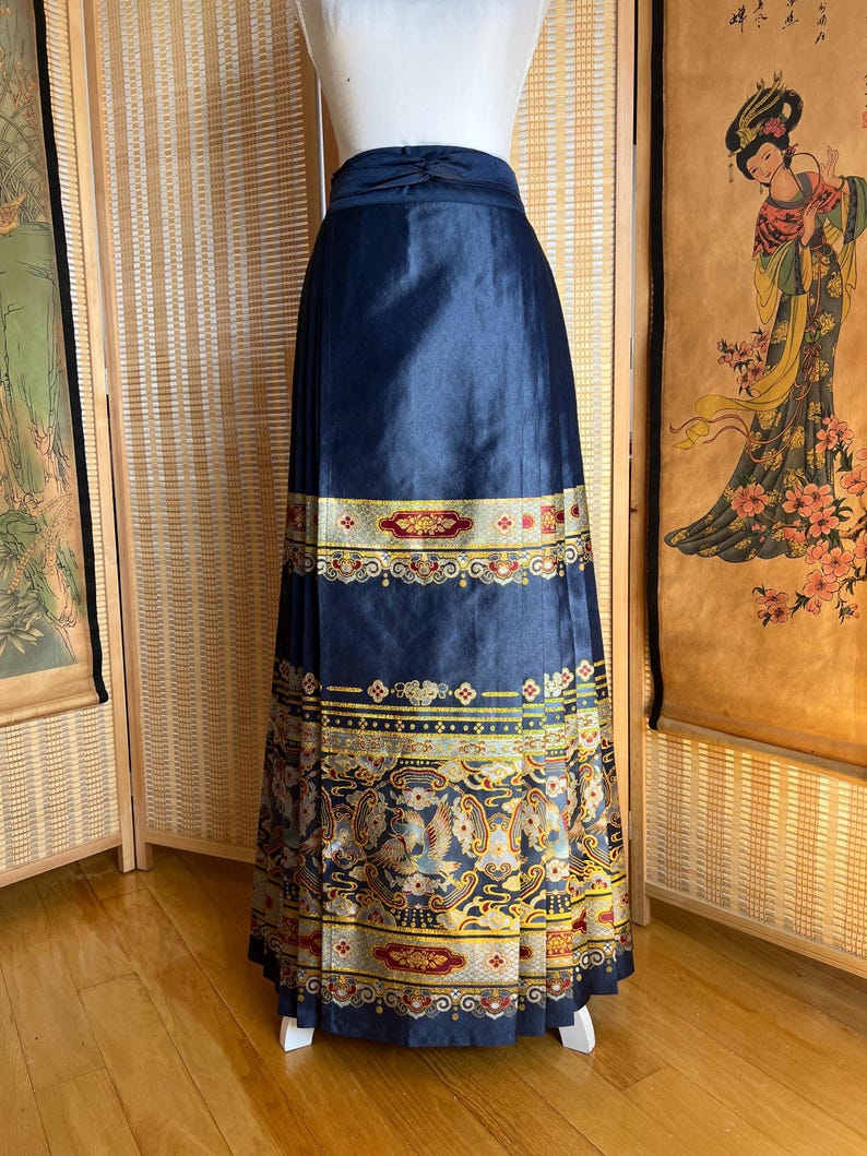 May include: A navy blue skirt with a decorative gold and red patterned border. The skirt is pleated and has a high waist. The lower border features a detailed design with birds and floral motifs.