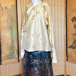 May include: A traditional Korean hanbok ensemble. The top is a pale yellow with floral patterns, a white collar, and long sleeves. The skirt is a vibrant blue with a colorful, ornate design. The outfit is displayed on a mannequin.