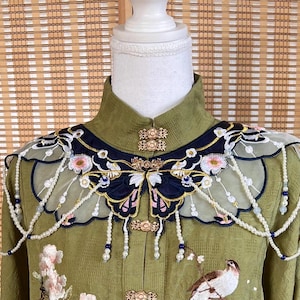 May include: Olive green embroidered top with a mandarin collar and ornate butterfly-shaped shoulder embellishments. The design features delicate pearl strands, floral accents, and a bird illustration. Gold-toned buttons and intricate detailing enhance the garment's elegance.
