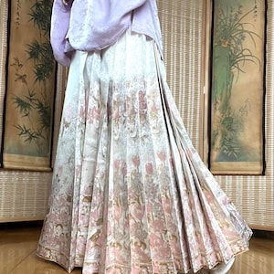 Pode incluir: Um traje tradicional com uma parte superior roxa clara e uma saia longa plissada. A saia tem uma base branca com um padrão floral em rosa e dourado. O traje provavelmente faz parte de um traje cultural ou histórico.