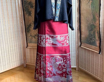 May include: A traditional outfit featuring a long, pleated red skirt with a detailed silver phoenix design. The outfit includes a black jacket with blue and white embroidered trim. The ensemble is displayed on a white surface.