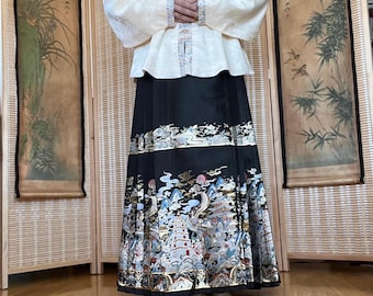 May include: A long-sleeved cream-colored top with a high collar and decorative trim paired with a black floor-length skirt. The skirt features a detailed, multi-colored embroidered pattern depicting traditional architectural structures, mountains, and clouds.
