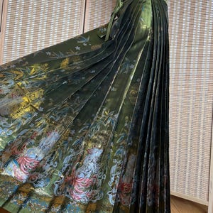 May include: Olive green pleated skirt with a decorative pattern featuring floral and animal motifs. The skirt has a high waist and a matching tie. The fabric has a metallic sheen, and the pleats are vertical.