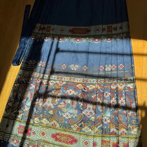 May include: A long, pleated skirt in shades of blue with an ornate, patterned design. The pattern includes birds, flowers, and paisley elements in red, gold, and white. The skirt has a matching blue tie.