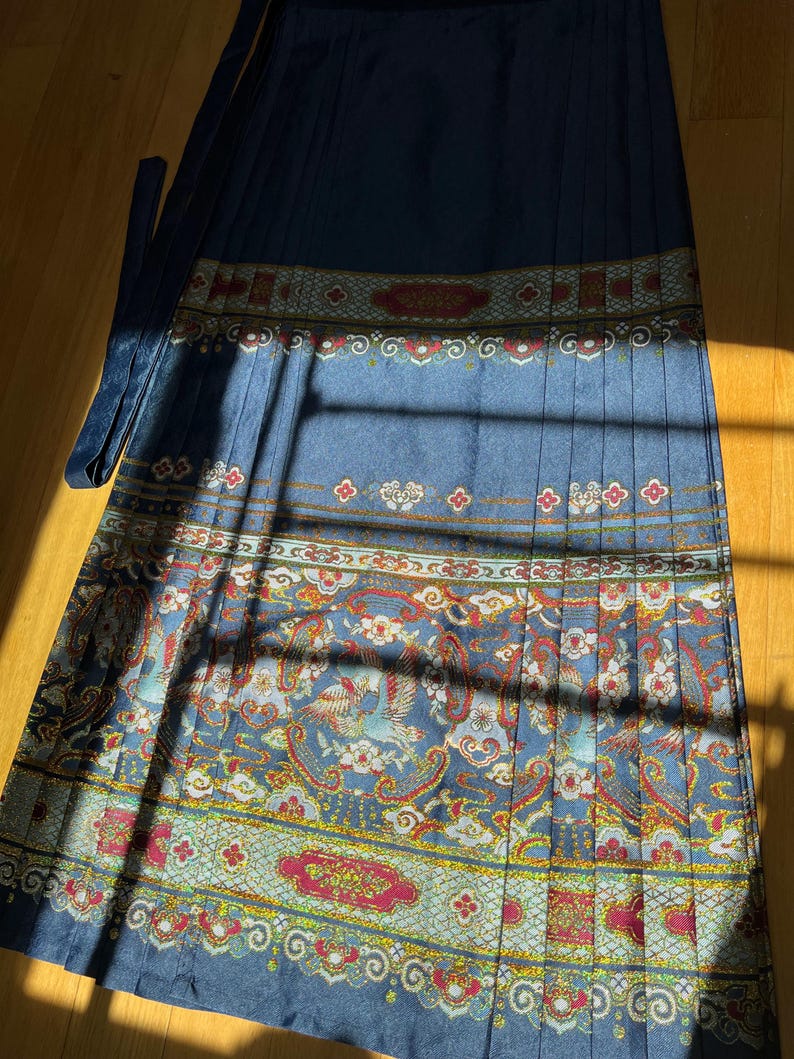 May include: A long, pleated skirt in shades of blue with an ornate, patterned design. The pattern includes birds, flowers, and paisley elements in red, gold, and white. The skirt has a matching blue tie.