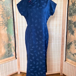 Könnte beinhalten: Ein dunkelblaues, wadenlanges Kleid mit Stehkragen, kurzen Ärmeln und einem dezenten, tonalen Blumenmuster. Das Kleidungsstück verfügt über einen Seitenschlitz und wird am Ausschnitt mit dekorativen Stoffknotenknöpfen geschlossen.