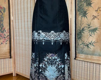 May include: A long, black skirt with intricate embroidery. The skirt features a detailed floral and mythical creature design in shades of silver, blue, and purple. The skirt is displayed on a white mannequin, showcasing its full length and design.