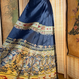 May include: A navy blue, pleated skirt with ornate, colourful embroidery. The skirt features a detailed pattern of birds, flowers, and geometric shapes in gold, red, and blue. The skirt is displayed on a mannequin.