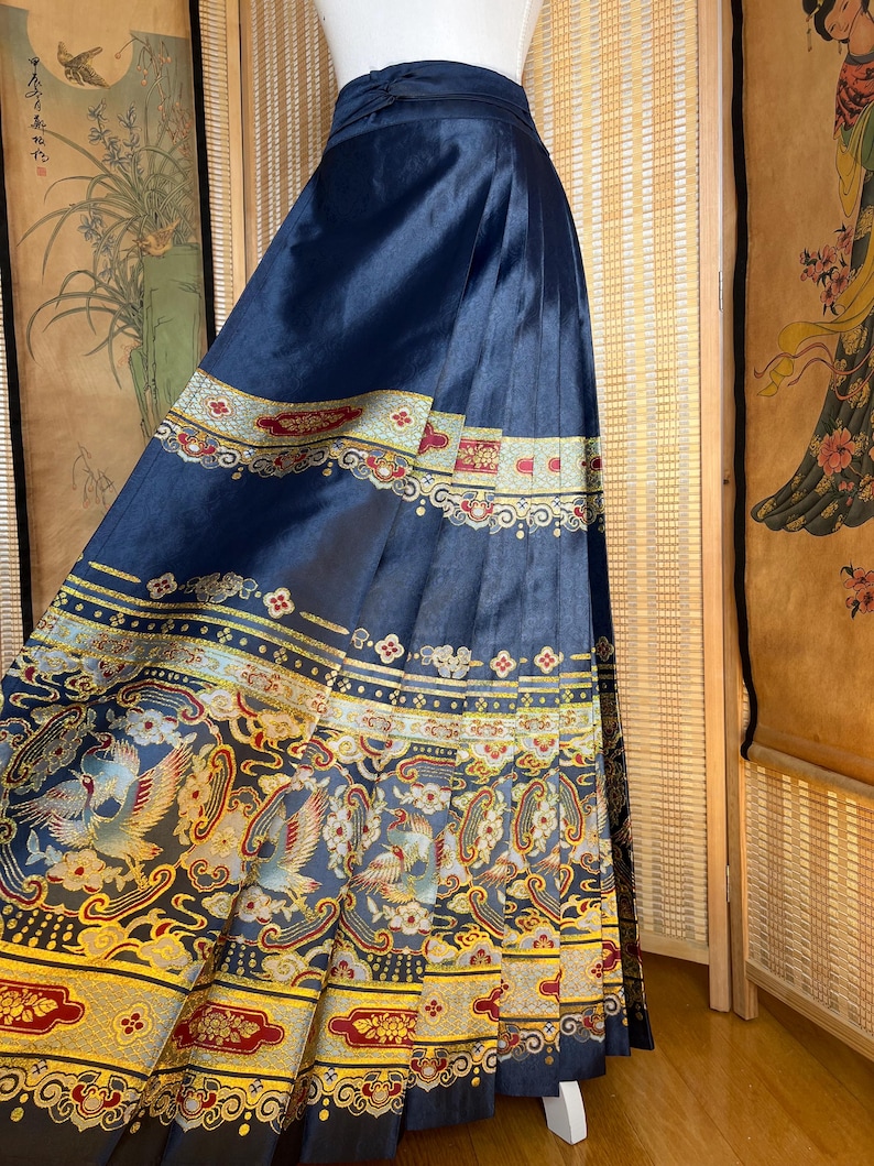 May include: A navy blue, pleated skirt with ornate, colourful embroidery. The skirt features a detailed pattern of birds, flowers, and geometric shapes in gold, red, and blue. The skirt is displayed on a mannequin.
