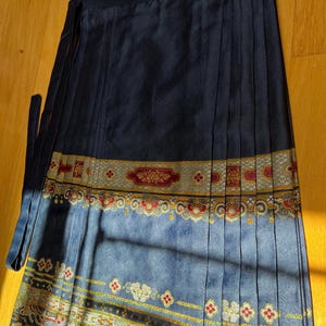 May include: A navy blue wrap skirt with a pleated design. The skirt has a decorative band with gold, red, and white patterns. It features long ties for closure and is made of a silky material.