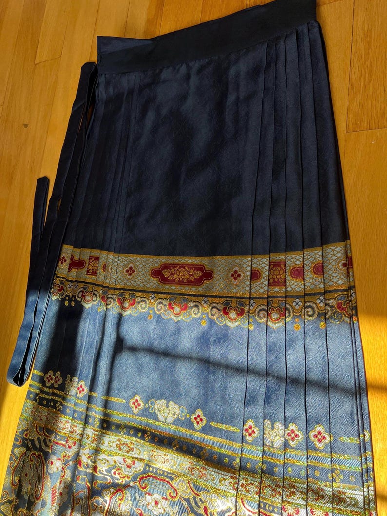 May include: A navy blue wrap skirt with a pleated design. The skirt has a decorative band with gold, red, and white patterns. It features long ties for closure and is made of a silky material.