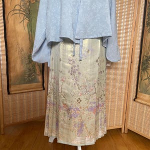 Pode incluir: Uma jaqueta azul clara texturizada sobre uma saia longa plissada. A saia apresenta um padrão floral em tons de roxo, rosa e dourado sobre um fundo verde claro. A roupa é exibida em um manequim branco.
