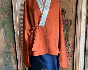May include: An orange wrap top with long sleeves and a floral pattern, paired with a navy blue skirt featuring a gold design. The top has a contrasting floral trim around the neckline and a tie closure.