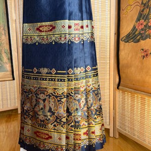 May include: A navy blue pleated skirt with ornate gold, red, and grey patterned bands. The skirt features detailed designs of birds and floral motifs, creating a rich, textured appearance. The skirt is displayed on a white stand.