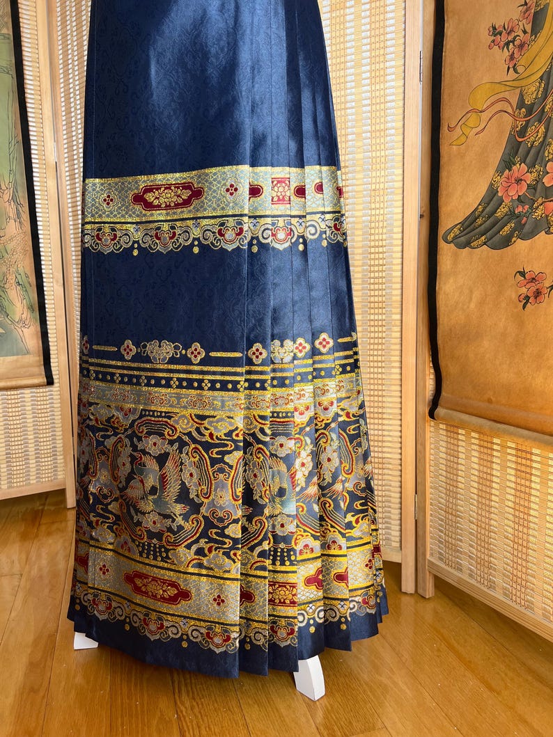 May include: A navy blue pleated skirt with ornate gold, red, and grey patterned bands. The skirt features detailed designs of birds and floral motifs, creating a rich, textured appearance. The skirt is displayed on a white stand.