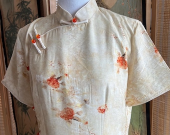 Puede incluir: Vestido color crema de manga corta con cuello mandarín y estampado floral. El vestido presenta patrones de flores rojas y naranjas y cierres decorativos de cuentas naranjas. El vestido se exhibe en un maniquí.