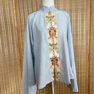 May include: A pale blue tunic top with long, wide sleeves. The front has a vertical panel with floral and bow designs in cream, red, and gold. It features a mandarin collar and decorative buttons. The sleeves have a decorative trim.