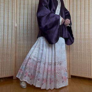 Pode incluir: Um traje tradicional japonês com um top de seda roxo escuro com mangas largas e uma saia branca com padrão floral. A saia tem um design plissado com detalhes de flores de cerejeira rosa. O traje é usado com sapatos prateados.