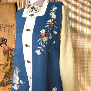 Puede incluir: Un atuendo tradicional de estilo chino con un chaleco azul con detalles blancos y bordados ornamentados de flores y frutas. El atuendo incluye una parte superior amarilla clara y un cuello lavanda, con botones decorativos.