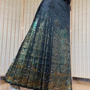 May include: A long, pleated skirt in dark teal with gold and green floral and bird-like patterns. The skirt has a wrap-around style with a black tie at the waist. The fabric has a subtle sheen, and the design is intricate and detailed.