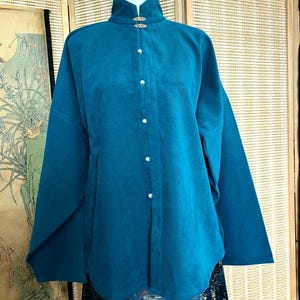 Peut inclure: Haut teal à manches longues avec un col mandarin et des boutons nacrés. Le haut présente des fermoirs décoratifs dorés au col et une coupe ample. Le vêtement est présenté sur un mannequin, mettant en valeur son design et son style.