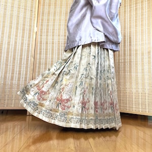 May include: A light purple silk top and a pleated skirt with a floral pattern. The skirt features a cream background with red and green floral designs and a blue border. The outfit is suitable for special occasions.