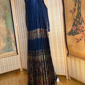 May include: A navy blue pleated skirt with a gold and red patterned border. The skirt has a wrap-around design with a matching tie. The skirt is displayed on a mannequin.