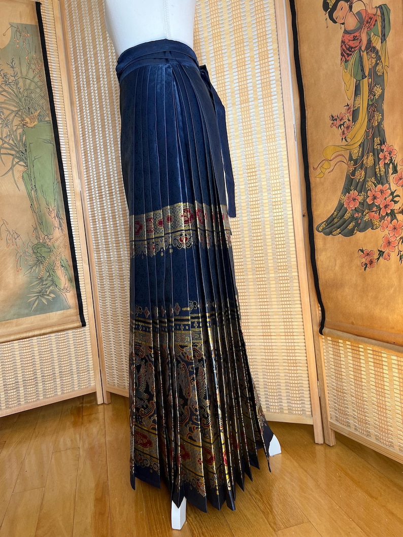 May include: A navy blue pleated skirt with a gold and red patterned border. The skirt has a wrap-around design with a matching tie. The skirt is displayed on a mannequin.