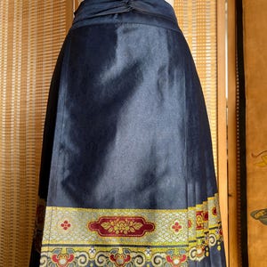 May include: A navy blue skirt with a decorative band at the hem. The band features a floral design in red, gold, and yellow. The skirt has a subtle pattern and a knotted detail at the waist.