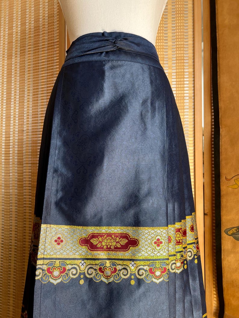 May include: A navy blue skirt with a decorative band at the hem. The band features a floral design in red, gold, and yellow. The skirt has a subtle pattern and a knotted detail at the waist.