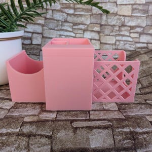 May include: A light pink desk organizer set consisting of three distinct sections. The central piece is a tall rectangular container, flanked by a curved open-top holder on the left and a square container with a lattice-style cutout pattern on the right.