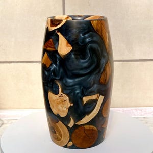 Peut inclure: Un vase en bois artisanal avec un intérieur en résine bleu foncé. Le vase est orné de morceaux de bois brun clair de différentes tailles, créant un design unique et artistique. Le vase cylindrique est posé sur une surface blanche.