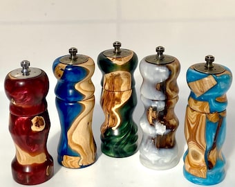 May include: Five cylindrical salt and pepper grinders made from a combination of natural wood and colorful resin. Each grinder features a metallic top mechanism and a unique, swirling pattern of wood grain mixed with red, blue, green, or white resin.